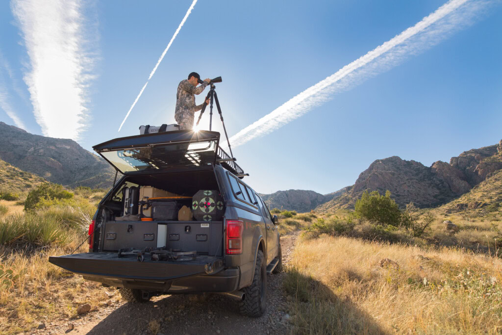 Big game hunter scopes out an area from on top of his truck. The truck bed is open showcasing an All-Weather TruckVault system